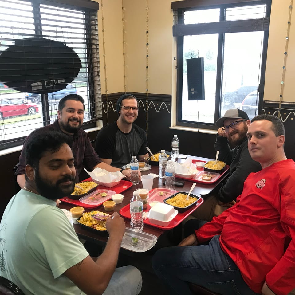 Six men smiling at a table, enjoying individual meals of rice and chicken in a casual restaurant.