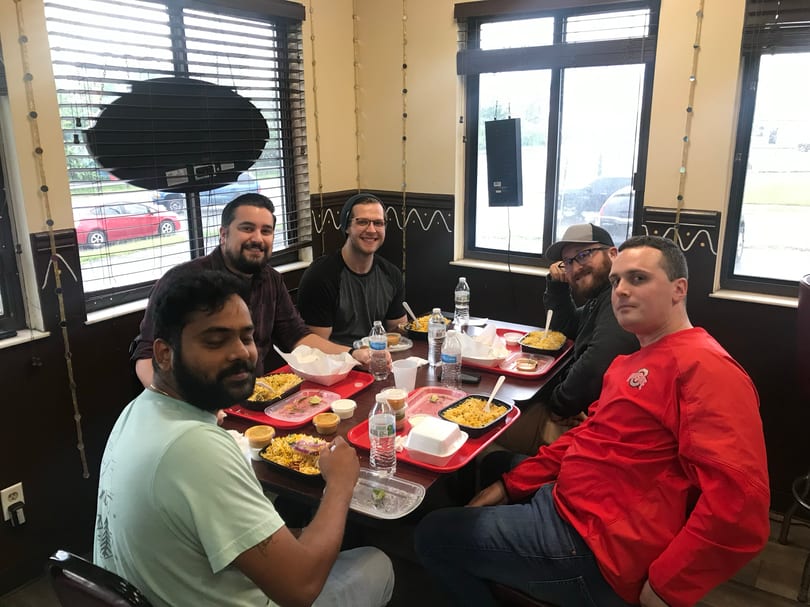 Six men smiling at a table, enjoying individual meals of rice and chicken in a casual restaurant.