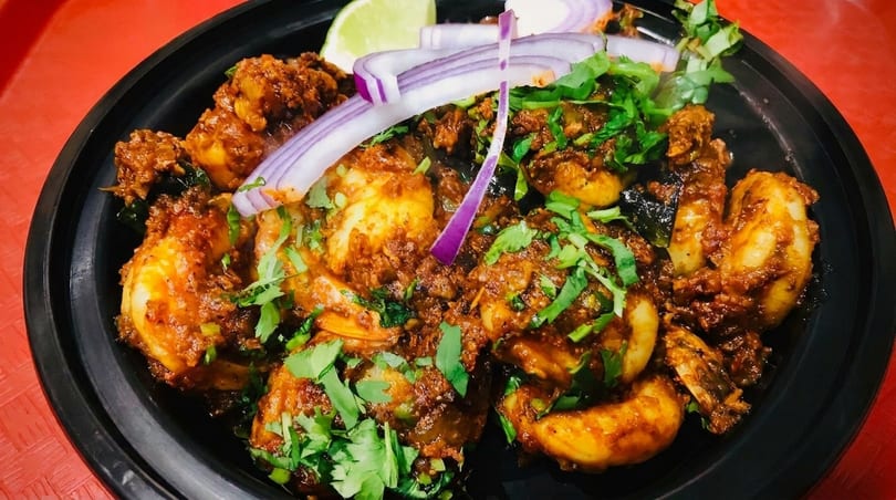 Spicy shrimp dish garnished with fresh cilantro, sliced red onions, and a lime wedge in a black bowl.