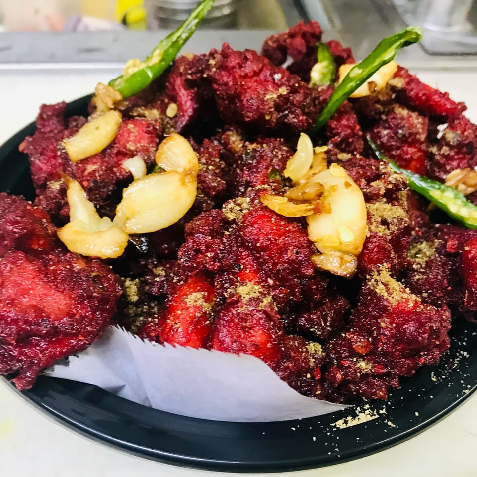 Deep red fried chicken pieces, garnished with garlic cloves and green chilies, served in a black bowl.