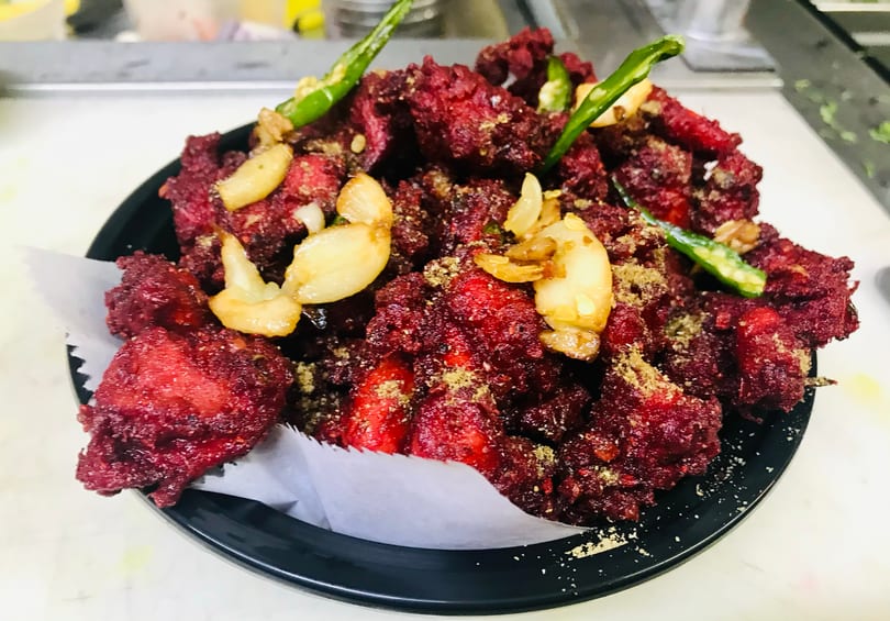 Deep red fried chicken pieces, garnished with garlic cloves and green chilies, served in a black bowl.