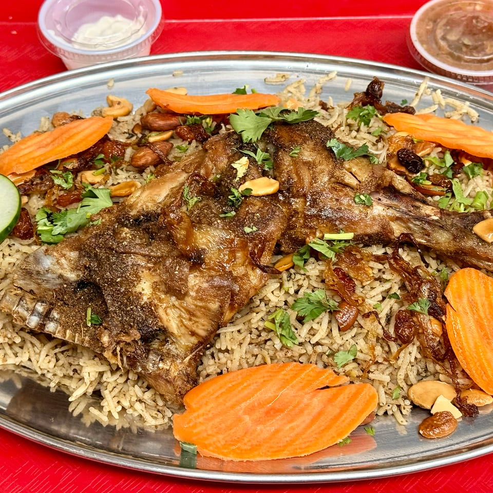 A large platter of Mandi rice with roasted lamb, garnished with carrots, cucumbers, nuts, and cilantro.