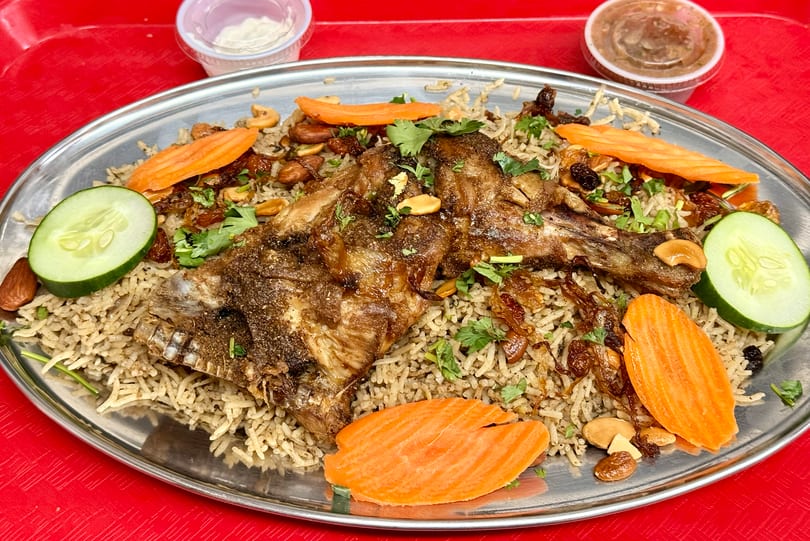 A large platter of Mandi rice with roasted lamb, garnished with carrots, cucumbers, nuts, and cilantro.