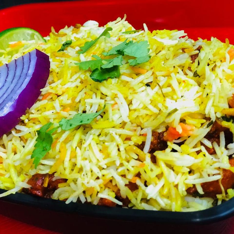 Chicken biryani with yellow rice, cilantro, red onion, and lime in a black bowl.