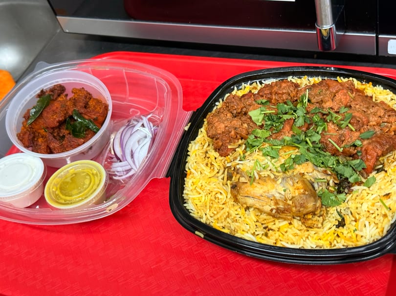 Chicken biryani with cilantro, a side of fried chicken, sliced onions, and sauces in takeout containers.