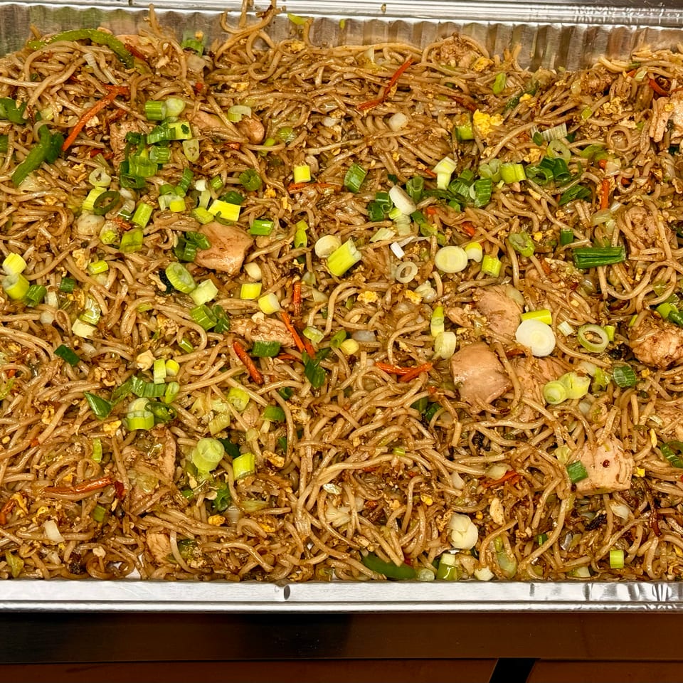 A large catering tray filled with chicken lo mein, garnished with green onions and carrots.