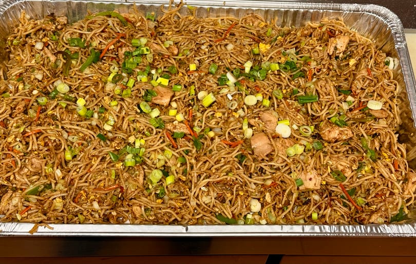 A large catering tray filled with chicken lo mein, garnished with green onions and carrots.