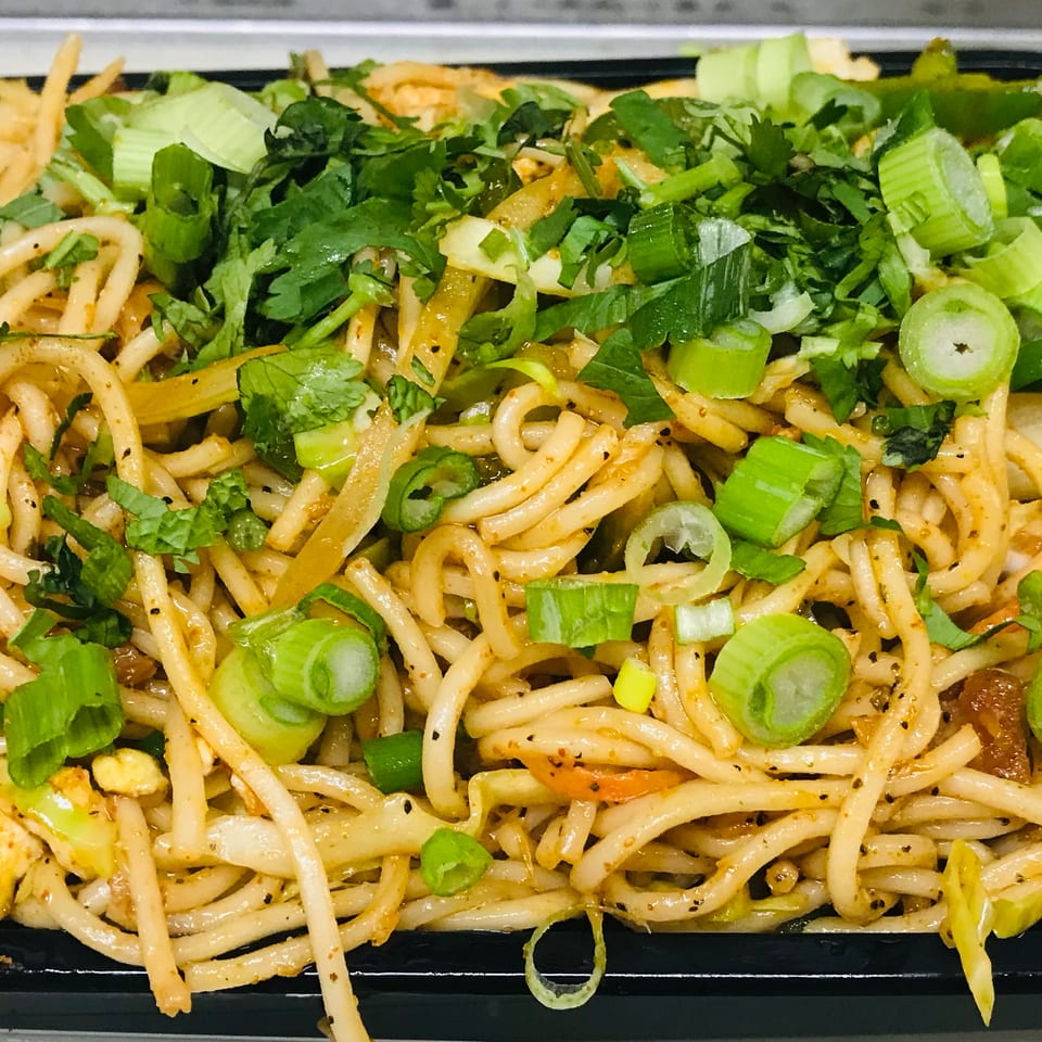 Close-up of a black container filled with noodles, chicken, and vegetables, topped with green onions and cilantro.