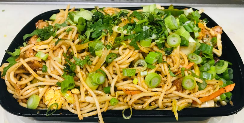 Close-up of a black container filled with noodles, chicken, and vegetables, topped with green onions and cilantro.