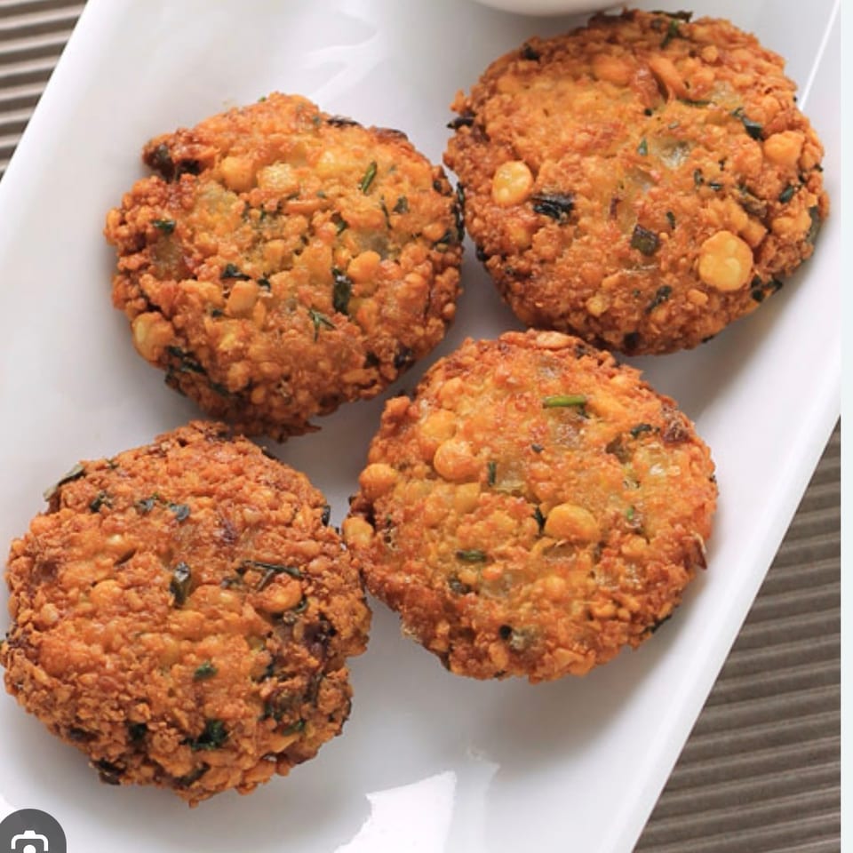 Four golden-brown, crispy lentil fritters (vada) with herbs on a white rectangular plate.