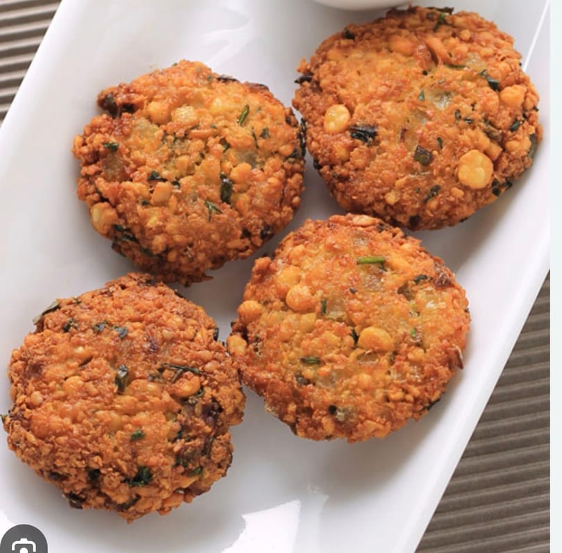 Four golden-brown, crispy lentil fritters (vada) with herbs on a white rectangular plate.