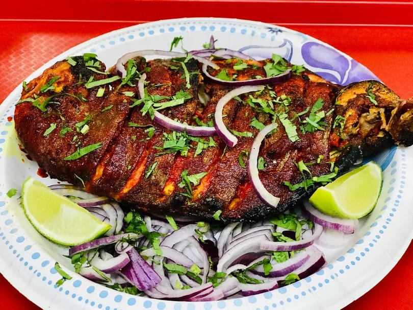 Whole grilled fish, garnished with red onion, cilantro, and lime wedges on a paper plate.