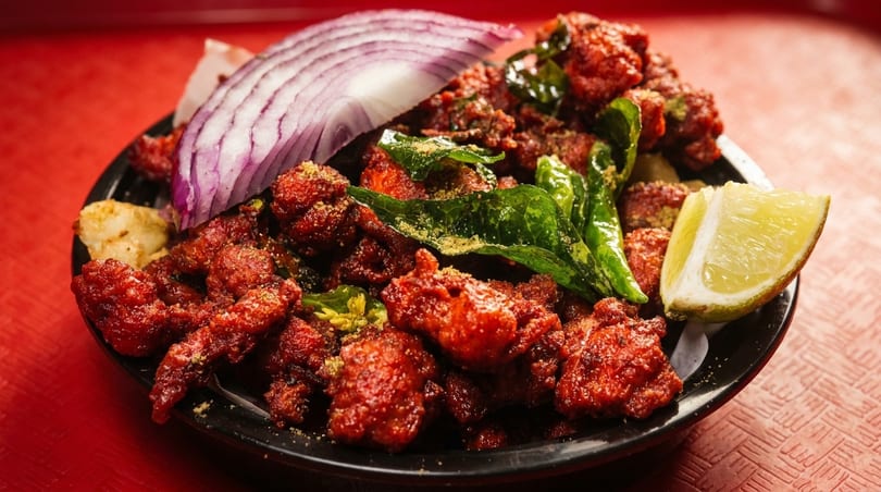 Spicy red chicken 65 with curry leaves, red onion, and a lime wedge on a black plate.