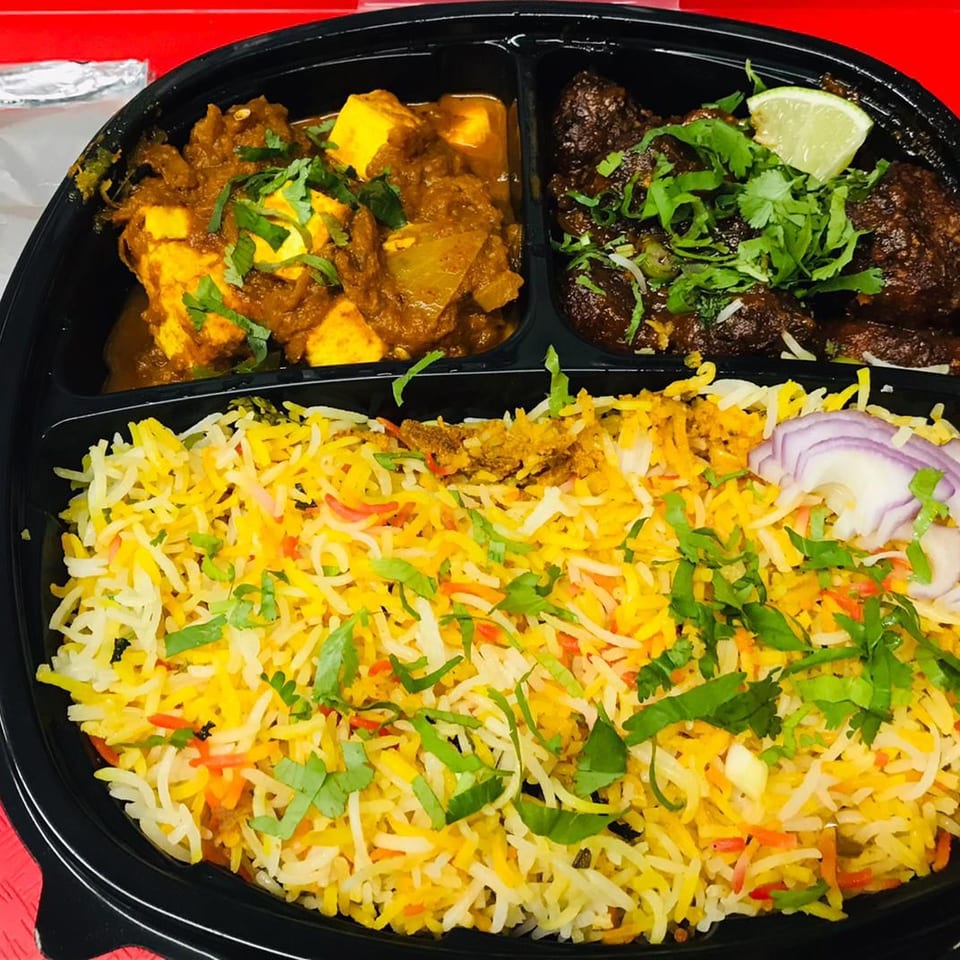 Indian meal with paneer curry, meat, biryani rice, and gulab jamun in a black to-go container.