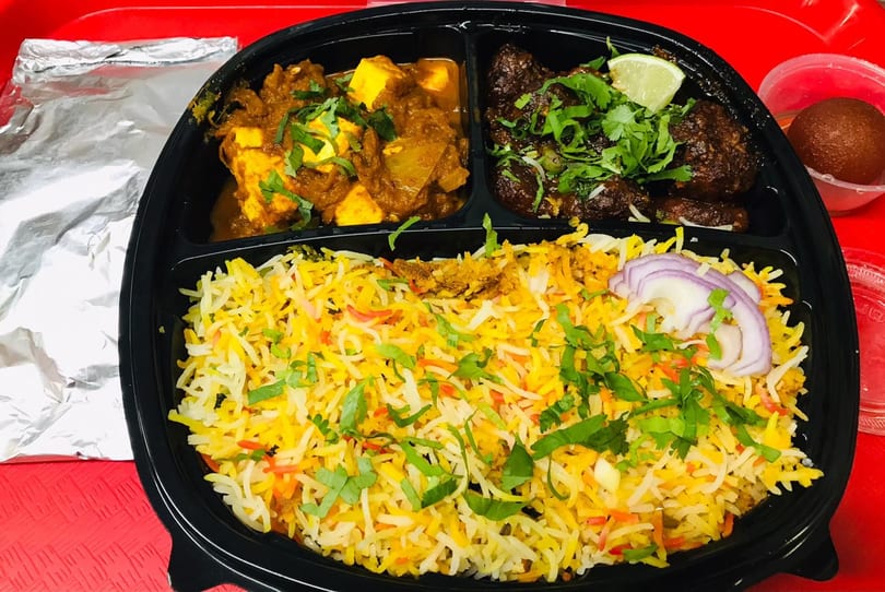 Indian meal with paneer curry, meat, biryani rice, and gulab jamun in a black to-go container.